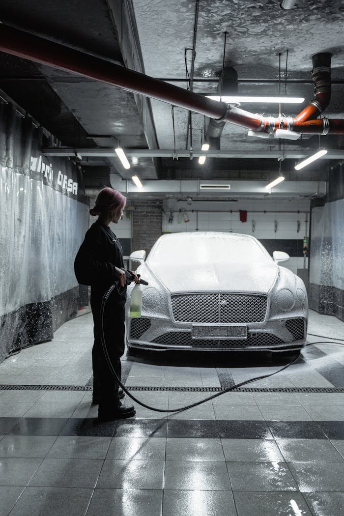 A woman uses a power hose to wash a luxury car with snow foam in a well-lit garage.